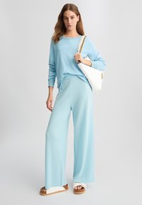 Maglione azzurro chiaro a maglia con polsini a coste, abbinato a pantaloni larghi coordinati. Borsa bianca con dettagli in catena dorata e sandali bianchi.