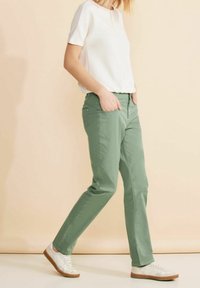 Henkilö, joka pukeutuu vaaleanvihreisiin housuihin, valkoiseen lyhythihaiseen paitaan ja valkoisiin tennareihin, kävelee kädet taskuissa beigeä taustaa vasten.