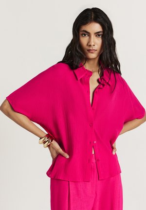 Chemise fuchsia à manches courtes, à coupe décontractée et en tissu texturé, associée à un pantalon coordonné et à des bracelets en or.