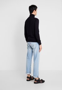 Sort turtleneck sweater, let strik, tætsiddende silhuet. Sammen med lyseblå rullede denim jeans og sorte sandaler. Ensfarvet baggrund.