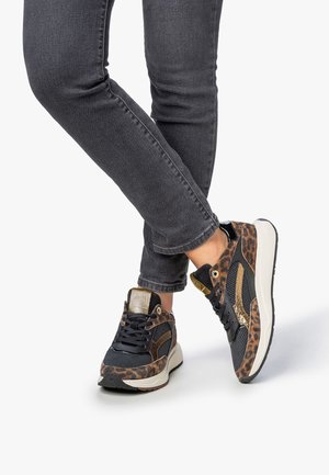 Persona che indossa jeans skinny grigi scuri e sneakers con stampa leopardata, dettagli neri e dorati, in piedi con le gambe incrociate.