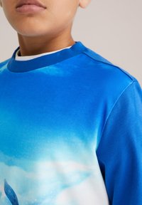 Mėlynas medvilnės džemperis su gradientu nuo šviesiai mėlynos iki tamsiai mėlynos, pasižymintis baltu apvaliu kaklu ir spausdintu dizainu, primenančiu vandenį.