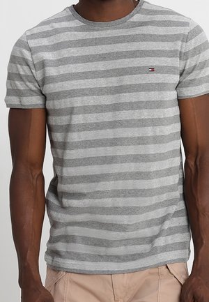 Mann trägt ein kurzärmeliges graues T-Shirt mit hellgrauen horizontalen Streifen und kleinem Logo auf der Brust sowie beige Hosen.