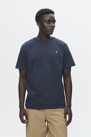T-shirt di cotone blu navy con maniche corte, scollatura rotonda e piccolo logo sul lato sinistro del petto. Design semplice, texture liscia. Indossato con pantaloni beige.