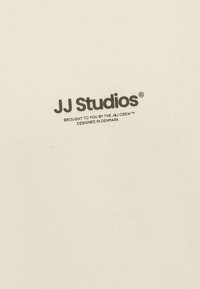 Pull-over crème clair avec texture côtelée. Présente le logo "JJ Studios" en gris foncé, avec un texte supplémentaire indiquant l'origine du design en plus petit.