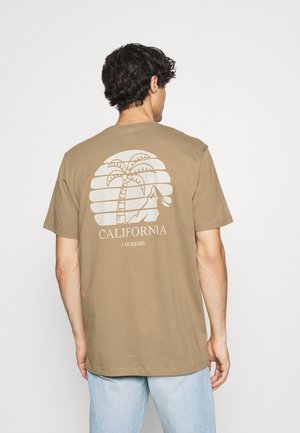 Homme portant un T-shirt marron clair avec un graphisme blanc représentant un palmier, une montagne, et le texte "CALIFORNIA / HOMME" au dos, assorti d'un jean bleu clair.