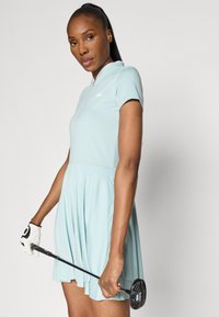 Svijetloplava golf haljina s ovratnikom na zatvarač, kratkim rukavima i mrežnim bočnim panelima. Model drži golf palicu noseći bijelu rukavicu.