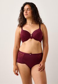 Nainen, jolla on kihara tumma hius, wearing matching burgundy pitsi rintaliivit ja korkeavyötäröiset alushousut, seisoo vaaleaa taustaa vasten.