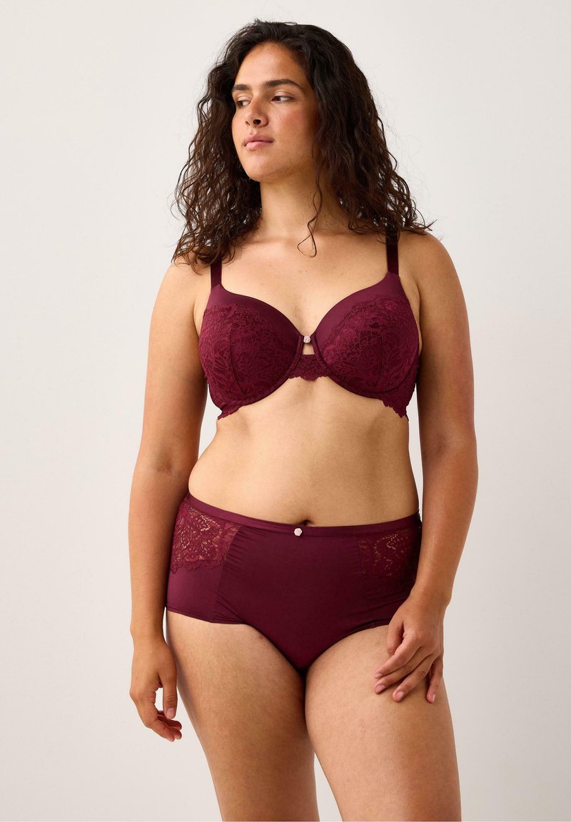 Nainen, jolla on kihara tumma hius, wearing matching burgundy pitsi rintaliivit ja korkeavyötäröiset alushousut, seisoo vaaleaa taustaa vasten.