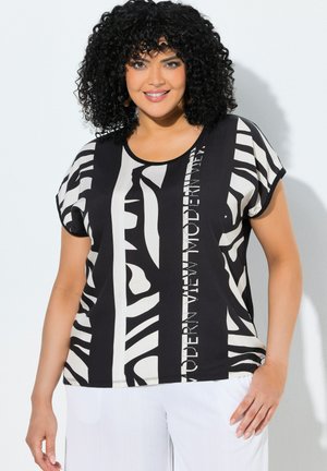 Frau mit lockigem dunklem Haar, die ein schwarz-weiß gestreiftes abstraktes Oberteil mit Text und weiße Hose trägt, lächelt vor hellem Hintergrund.