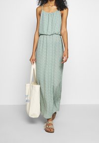Robe maxi verte sans manches avec une taille froncée, présentant un subtil motif de petits pois blancs ; assortie avec des sandales marron et un sac fourre-tout crème.