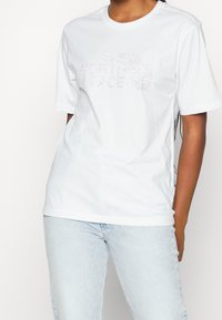 Bílý bavlněný tričko s krátkým rukávem, které má na hrudi lehký grafický logo "The North Face", v kombinaci s světle modrými džíny.