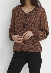 Blusa marrone a fantasia con scollatura a V, bordi neri e dettagli con frange. Maniche lunghe con polsini elastici, tessuto fluido e stampa floreale.