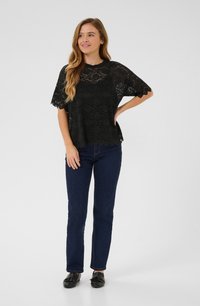 Svart spetsblus med korta ärmar och scallopad nederkant, bärs över mörkblå raka jeans och svarta skor. Slät textur, blommönster.