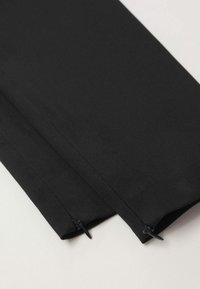 Gros plan sur des jambes de pantalon en tissu noir avec de petites fermetures éclair noires aux ourlets sur une surface claire.