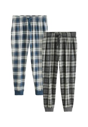 Tetoválásokkal rendelkező férfi áll mezítláb, kék-fehér kockás pizsamanadrágban egy bézs és szürke falú hálószobában.