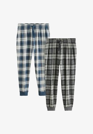 Tetoválásokkal rendelkező férfi áll mezítláb, kék-fehér kockás pizsamanadrágban egy bézs és szürke falú hálószobában.