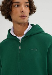 Sudadera verde con cremallera hecha de tela suave, con capucha, cuello blanco y un logo embosado en el pecho. Hardware de cremallera de alta calidad visible.