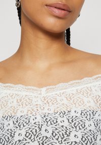 Balta off-the-shoulder nėriniuota palaidinė su juodais gėlių raštais, turinti žakardo nėrinių apvadą ir skaidrų tekstūrą.
