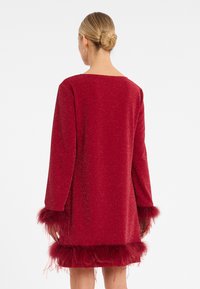 Rotes Glitzerkleid mit langen Ärmeln, verziert mit Federakzenten an den Bündchen und dem Saum. Glatte Textur mit einer lockeren Passform und weitem Ausschnitt.