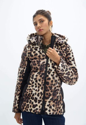 Joven mujer con chaqueta acolchada de estampado de leopardo con capucha, sosteniendo la cremallera, de pie frente a un fondo blanco liso.