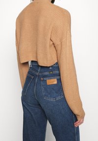Krátký žebrovaný svetr ve světle hnědé barvě s vysokým límcem; zkombinován s vysokým pasem tmavě modrých džín s koženou značkou Wrangler.