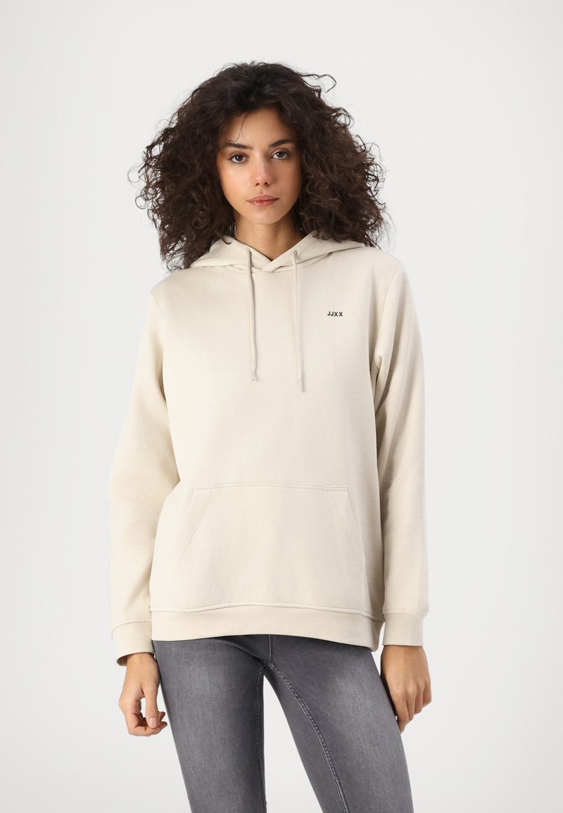 Beige kokvilnas hoodie ar priekšējo kabatu, kapuci ar auklu un mazu melnu logotipu kreisajā krūtīs, kombinēts ar pelēkiem džinsiem.