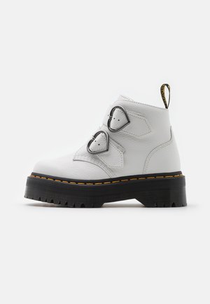 Bota blanca de cuero hasta el tobillo con suela negra gruesa, costuras amarillas y dos hebillas metálicas en forma de corazón en el lateral.
