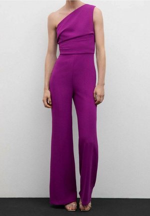 Henkilö, jolla on päällään hihaton, yhden olan magentanvärinen haalarihousupuku leveälahkeisilla housuilla ja remmikkäillä avokkaille avoimilla korkeakorkoisilla kengillä yksiväristä taustaa vasten.