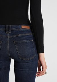 Person med mörkblå denimjeans med brun etikett och svart långärmad topp, hand avslappnat vid sidan mot vit bakgrund.