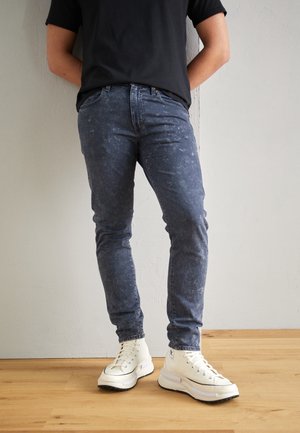 Personne portant un jean bleu foncé délavé, des baskets montantes blanches et un t-shirt noir, debout sur un sol en bois.