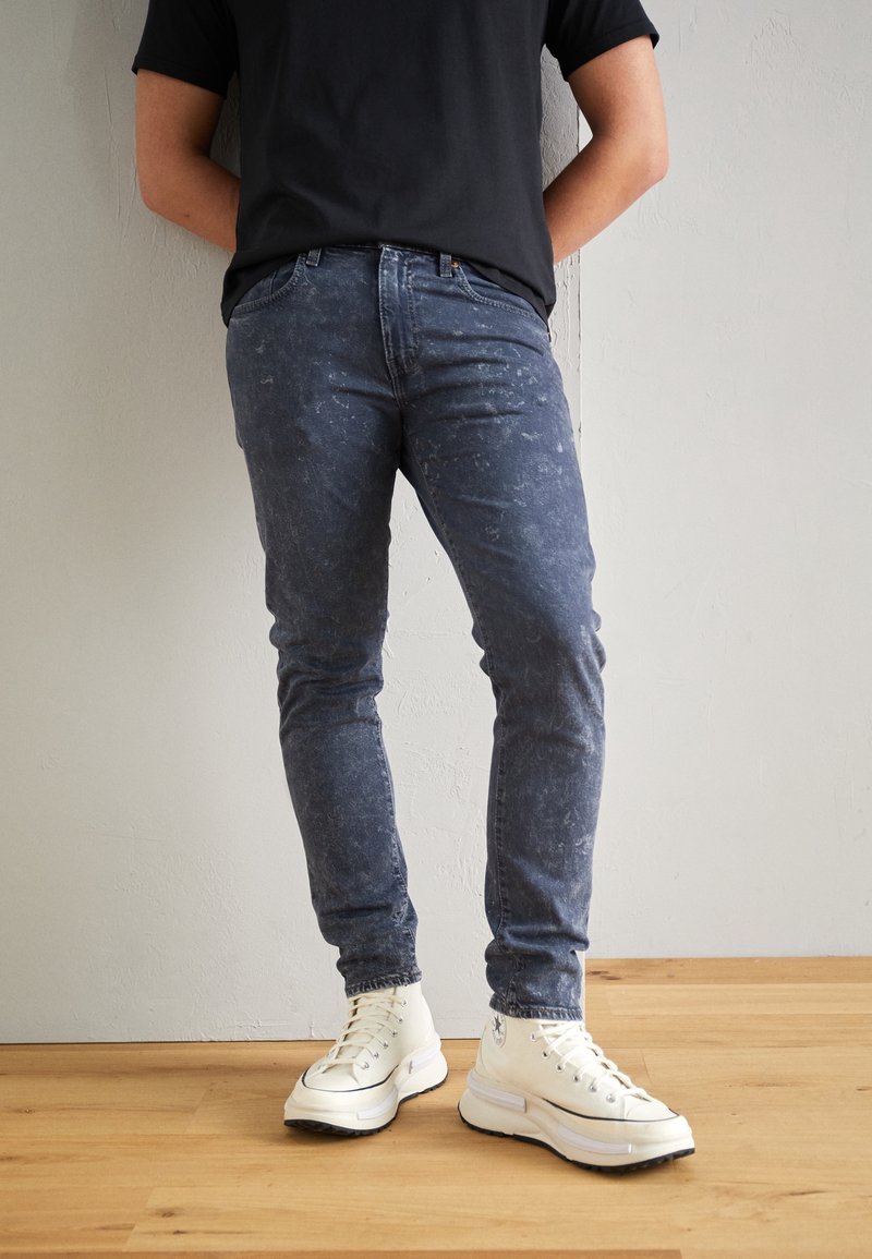 Personne portant un jean bleu foncé délavé, des baskets montantes blanches et un t-shirt noir, debout sur un sol en bois.