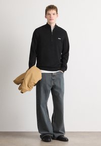 Μαύρο πουλόβερ με φερμουάρ και μικρό λευκό λογότυπο, συνδυασμένο με φαρδυά μπλε τζιν και κίτρινη μαστίχα ζακέτα κρεμασμένη στον βραχίωνα.