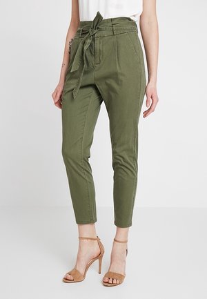 Vrouw draagt olijfgroene paperbag taillebroek met gestrikte riem en beige high-heeled sandalen, staand tegen een effen achtergrond.