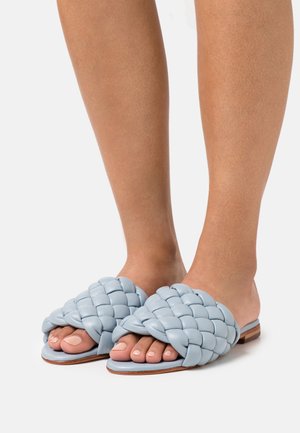 Pieds portant des sandales en cuir tressé bleu pâle à enfiler, avec des orteils ouverts et des semelles plates en bois sur fond blanc.