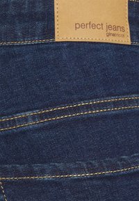 Tmav�ě modré džíny vyrobené z denimu s kontrastním zlatým šitím a obdélníkovou hnědou koženou nášivkou s nápisem "perfektní džíny giatritcot."