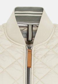 Beige gesteppte Jacke mit hohem Kragen, grau gestreiftem Rippstrickbesatz und einem Frontreißverschluss mit einem braunen Leder-Ziehband.