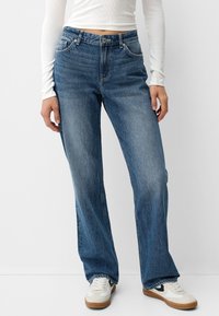 Persona que lleva puesto jeans rectos azules, una camiseta blanca de manga larga y zapatillas blancas con detalles en negro y suela de goma.