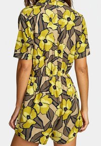 Combinaison fleurie avec des manches courtes, taille élastique, et un fond beige orné de grandes fleurs jaunes et d'accents sombres.