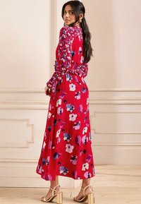 Vestido floral rojo con mangas largas y abullonadas y cintura ajustada. Presenta varias flores de diferentes colores sobre un fondo brillante. Sandalias doradas de tacón.