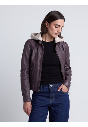 Kvinde med kort mørkt hår iført en mørkelilla læderjakke med beige hætte, sort top og blå jeans, kigger ned med hånden i lommen.