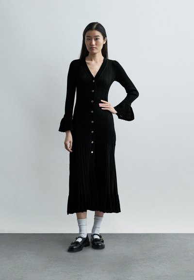 Γυναίκα στέκεται με το ένα χέρι στη μέση, φορώντας μακρύ μαύρο φόρεμα με κουμπιά και καμπανούς στα μανίκια, λευκές κάλτσες και μαύρα μοκασίνια πάνω σε γκρι πάτωμα.