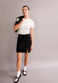 Witte t-shirt met klein logo, zwarte mini-jurk met een zijsplit en zwarte schoenen met witte accenten. Model houdt een zwarte blazer vast.
