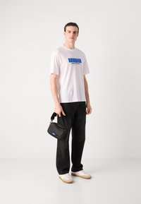 Camiseta blanca con logo azul "HUGO" y texto "CREATIVE DEPT.", combinada con jeans negros y un pequeño bolso negro tipo bandolera. Zapatillas blancas con suelas beige.