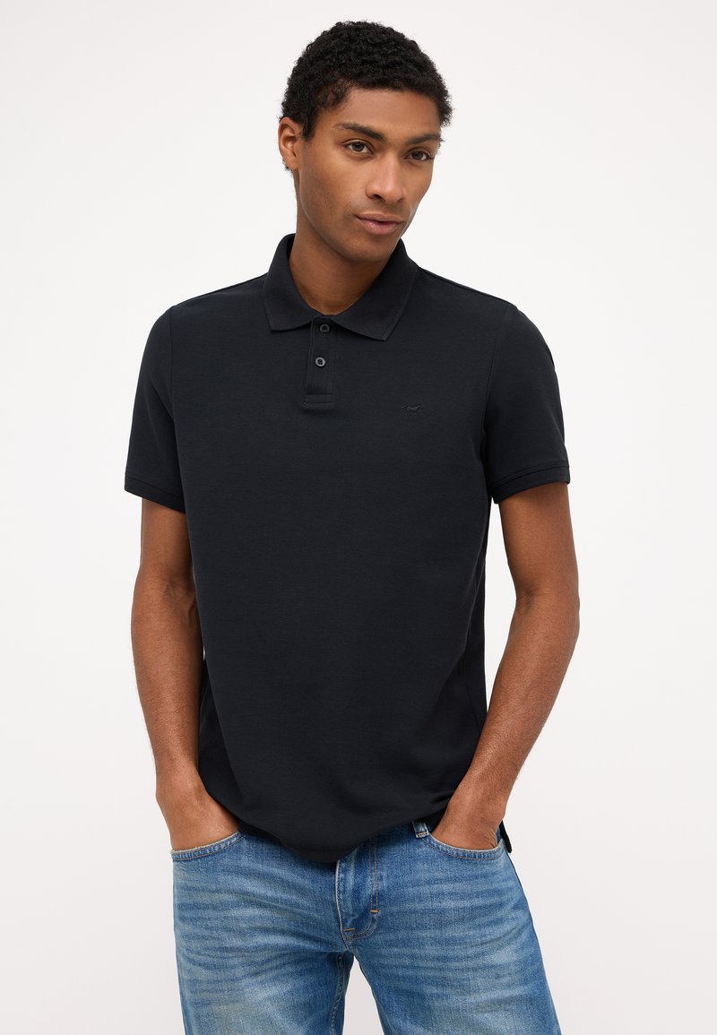 Polo negro de manga corta, con tapeta de tres botones y un logo sutil. Combinado con jeans azul claro, mostrando un ajuste casual.