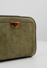 Borsa in suede verde con forma rettangolare, caratterizzata da un lucchetto ambra lucido e bordi neri. Texture liscia e dettagli di design minimalisti.