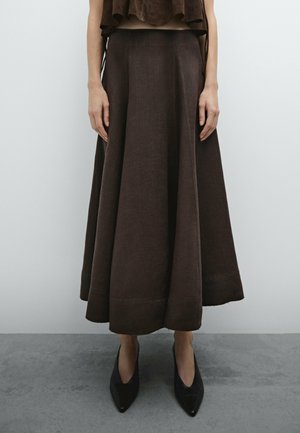 Femme portant une longue jupe évasée marron foncé avec des escarpins noirs à bout pointu, se tenant contre un mur clair et un sol gris.