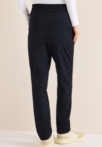 Pantalon de jogging gris foncé avec une texture douce, une ceinture élastique et deux poches arrière. Associé à des baskets de couleur claire.