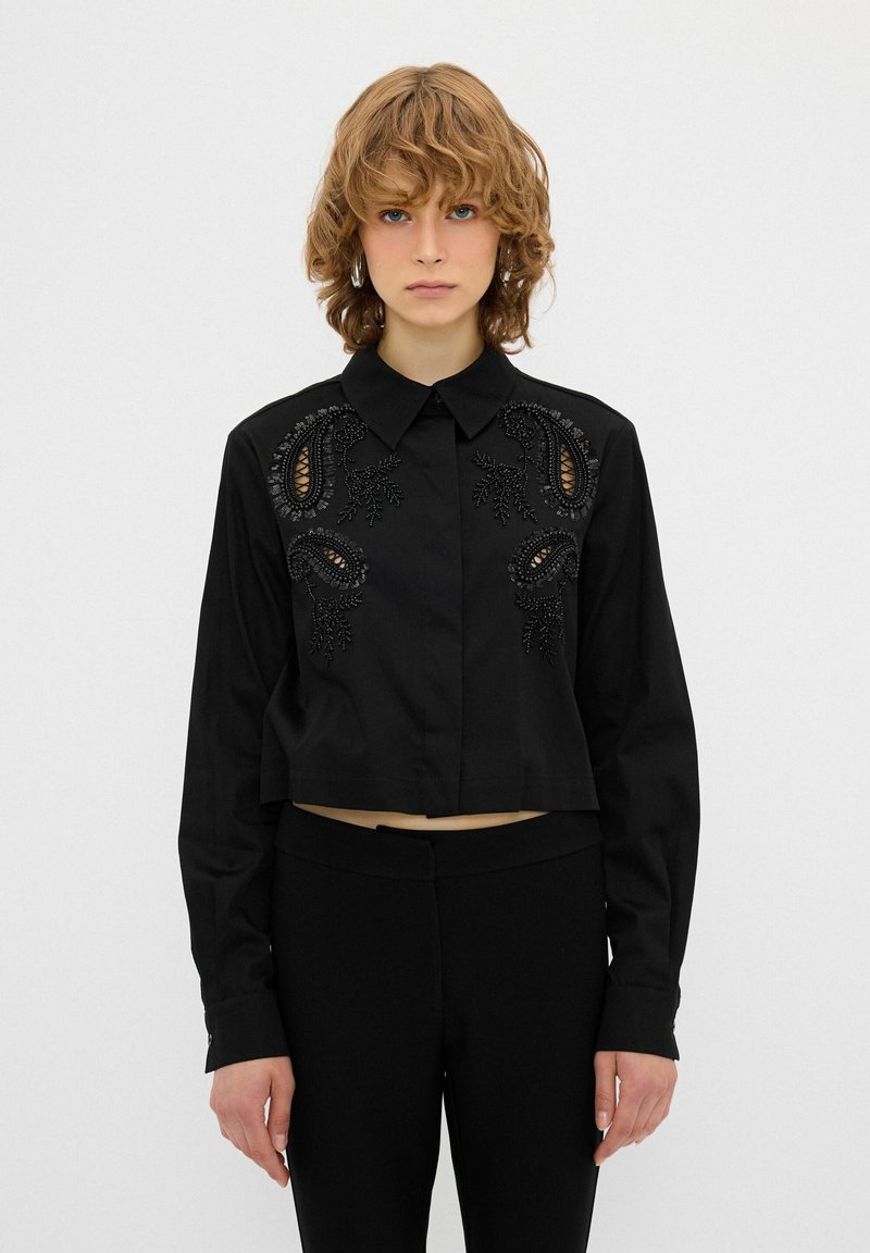 Schwarzes, kurz geschnittenes Shirt mit langen Ärmeln, das aufwendige schwarze Stickereien und ausgeschnittene Details auf der Brust aufweist. Klassisches Kragen-Design.