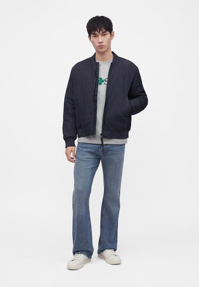 Jeune homme debout portant une veste bomber noire, un sweat-shirt gris à motif, un jean bleu et des baskets blanches sur un fond blanc uni.